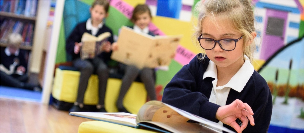 Young girl with glasses reading a book in a classroom, with three other children reading in the background.